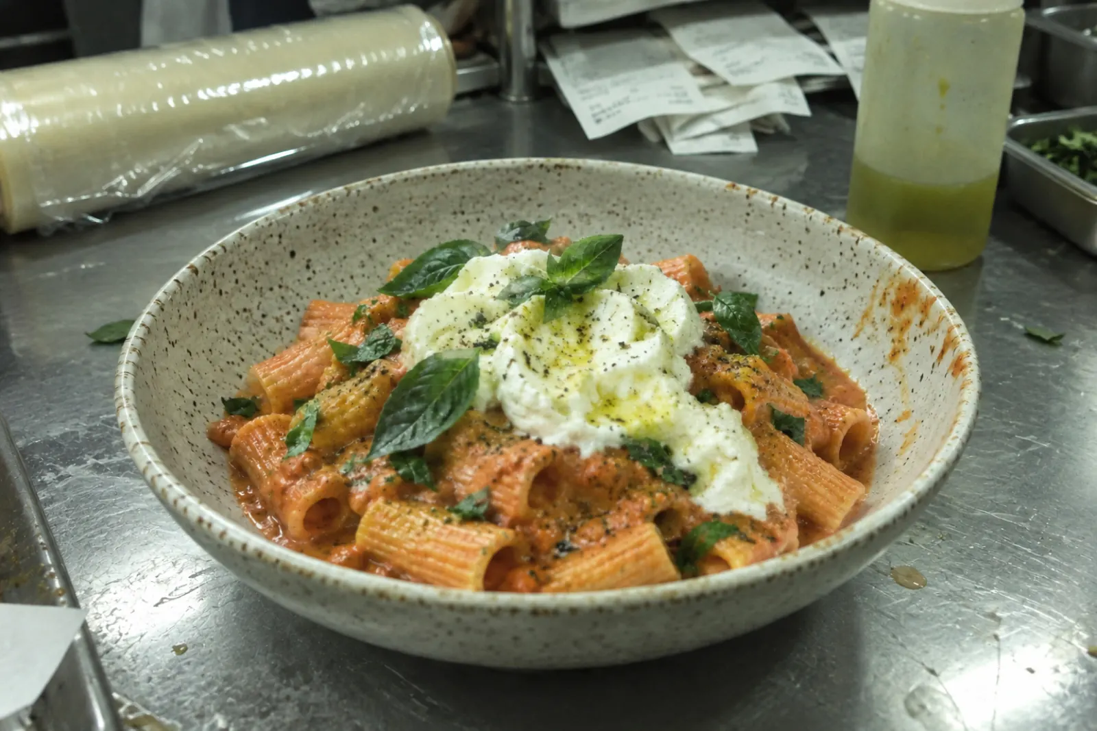 A dull, green-tinted phone photo of rigatoni on a cluttered steel prep counter